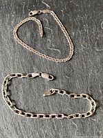 Two sterling silver bracelets on textured dark stone background; one with rectangular cable chain links and the other with marine/anchor chain links, both with lobster clasps.