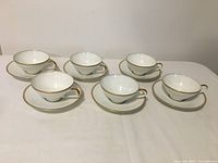 Six white porcelain cups and saucers with gold trim set on a white surface, arranged in two rows.