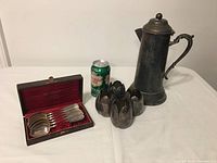 Wide shot of the boxed tablespoons, votive holders fashioned from tablespoons, and tall metal jug with dent and detached hinge