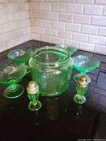 Photo showing the full set including jar, four stemmed glasses, and salt and pepper shakers on a black countertop near white brick wall.