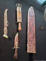 Four antique blades and their sheaths displayed on a black surface showing the variety in size, handle materials, and sheath decoration.