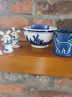 Full view of all items including two small windmill figurines, a ceramic bowl, and a blue pot with cow embossing on a wooden shelf.