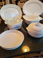 Wide shot of complete set showing white ceramic casseroles including Emile Henri and Corningware pieces, glass lids, and two white serving platters.