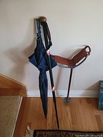 Photo of a golf lot showing a folded dark blue umbrella with wooden handle, a black walking stick with wooden knob handle, and a portable shooting stick seat with brown leather seat on wooden floor.