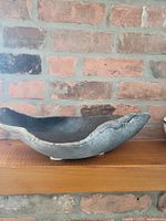 Side profile of a handmade graphite grey bonsai bowl showing natural irregular edges and a textured surface, set against a brick wall background.