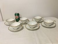 Set of 6 white porcelain teacups each placed on matching saucers with a soda can for size comparison on a white cloth background.