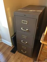 Front view of brown metal filing cabinet showing three drawers with label holders and handles.