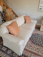 Side perspective of cream sofa with two peach throw pillows on patterned rug near wooden staircase.