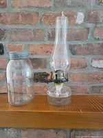 Photo showing the Queen Mary oil lamp with clear glass chimney and decorative embossed glass base next to the Crown glass jar with metal lid on a wooden surface against brick wall.