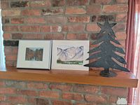 Photo showing two framed Elizabeth Gibson artworks and metal Christmas tree sculpture on wooden mantle