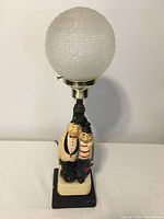 Front view of vintage lamp with Laurel and Hardy style figurine base and textured white glass globe shade. Shows figurine detail and paint condition.