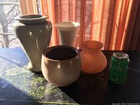 Five assorted vases on a surface with orange curtains in background, accompanied by a soda can for scale.