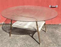 Side view of the round 36" textured glass patio table with bronze legs and woven vinyl shelf under base, showing wear and paint loss on legs.