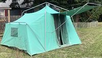 Full view of green vintage canvas tent with metal frame and peaked roof, front entrance flaps open, showing tent structure and material condition