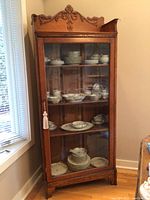 Front view of wooden curio cabinet filled with china dishes, closed glass door, carved details at top, tassel on door handle