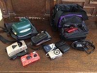 Various used cameras including Samsung Maxima Zoom Evoca and Konica, along with camera bags and accessories on wooden surface