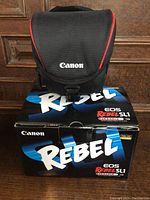 Canon branded black carrying case resting on top of the original Canon Rebel EOS Rebel SL1 box. The box shows the branding and Full HD video feature.