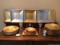 Front view of stack of square metallic chargers in silver and gold tones on a wooden sideboard, with clear glass bowls and etched glasses in front.