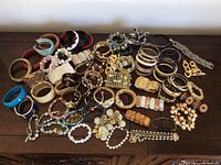 Photo showing a large assortment of various costume jewelry bracelets arranged on a wooden surface including bangles, beaded bracelets, chain links, and stretch styles in multiple colors.