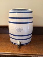 Full front view of the ceramic crock showing blue horizontal stripes and metal spigot at base.