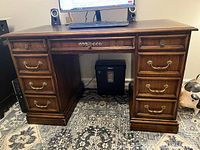 Full front view of a rectangular wooden pedestal desk with multiple drawers, brass handles, and a polished finish placed on a patterned carpet.