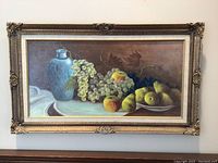 Wide-angle photo showing entire framed still life painting with blue vase, grapes, peaches, and pears on a table inside an ornate frame with minor scratches and dust.