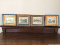 Four framed landscape prints lined up on a wooden shelf, mostly gold frames with blue inner border.