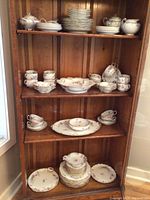 Full set of porcelain dinnerware displayed on wooden shelf, showing multiple pieces including plates, cups, bowls, and covered sugar bowls with floral and gold trim design.