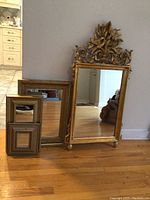 Four gilt framed wall mirrors of various sizes leaning against wall on hardwood floor