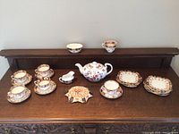 Full view of all china pieces displayed on wooden furniture surface including teapot, cups, saucers, plates, cream pitcher, and serving dish.