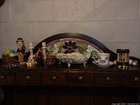 Wide view of all items on a dark wooden surface with stained glass background (not included). Items include a brown boot planter, caricature man figurine, porcelain cherubs, teacup with saucer, small teapot, and other small decorative figurines.
