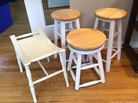 Five seating items including four wooden stools with light wood round tops and white legs, plus one white folding chair with slatted seat, all against a hardwood floor background.