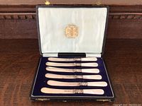 Full view of the open presentation case showing 8 butter knives with mother of pearl handles arranged neatly on blue velvet lining, white satin on the lid with gold crest emblem.