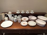 Wide view of mixed porcelain dinnerware and serveware including cups, mugs, plates, and oval serving dishes arranged on a wooden table.