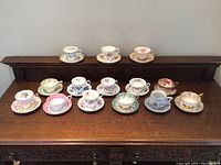 Front view with all ten tea cups and saucers displayed in two rows on a wooden surface, showing variety in floral and design patterns.