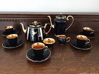 Photo showing the entire black and gold Wade England China tea set including two teapots, six cups, five saucers, creamer, and sugar bowl arranged on a wooden surface.