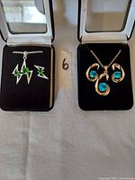 Two necklace and earring sets displayed in open boxes; left set is silver-tone with green gemstones, right set is gold-tone with blue gemstones.