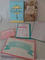 Set of bridal shower party games and books laid out on a white surface, showing the spiral-bound book, the Carolyn Cooper book, and five loose game sheets.