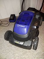 Front three-quarter view of mower on carpet with battery and charger beside it