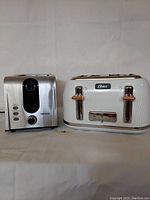 Two toasters displayed side by side on neutral fabric background; Heritage 2-slice toaster in steel with black details, Oster 4-slice toaster in white with gold accents