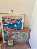 Three framed art pieces leaning against a white wall including a large colorful seaside scene with sailboats, a vintage world map, and a small vibrant floral print.