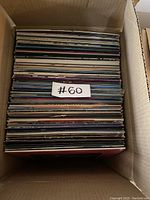 Top-down view of open cardboard box filled with vinyl records, approximately 60, sleeves showing various colors and designs.