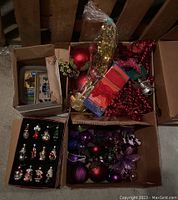 Top-down view of four cardboard boxes filled with assorted Christmas decorations including baubles, floral picks, and boxed ornaments