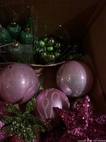 Box of mostly pink and a few green Christmas baubles and glittery green leaves and a purple glitter star ornament.