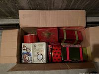 Photo of open cardboard box containing an assortment of Christmas-themed tins and boxes in red and green hues.