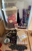 Photo showing clear storage bins filled with wrapping paper, gift bags, a purple decorative tree, colorful round ornaments in tubes, decorative wreaths, and beaded garlands.