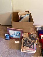Photo showing the cardboard box filled with assorted car magazines, framed photos, and other magazines/books spread on the floor.