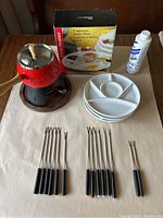 Photo of all items in the lot including fondue pot on wood base, 7 stoneware fondue plates, 14 fondue skewers with black handles, and boxed stoneware plates.