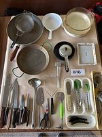Photo of various kitchen utensils, cast iron skillet with lid, Pyrex glass bowls, CorningWare cookware, strainer, ladle, spatula, tongs, peelers, scissors, and potato masher