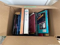 Open cardboard box filled with various books of different sizes and subjects including cookbooks, architecture, and novels.
